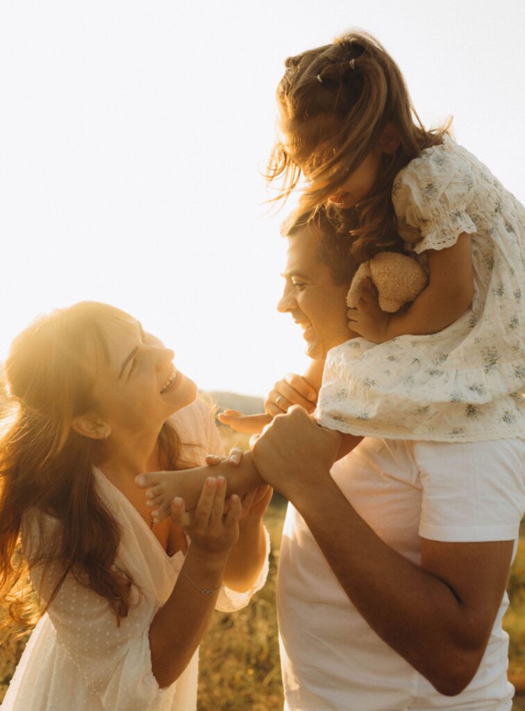 Eine glückliche Familie im Gegenlicht der Abendsonne: Der Vater trägt die kleine Tochter auf den Schultern, während die Mutter daneben steht und beide lachend ansieht.