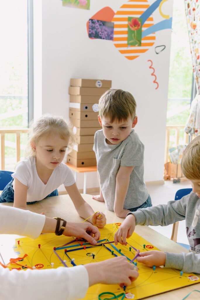 pexels-kseniachernaya-8535146 Drei Kinder sitzen und stehen konzentriert um einen Tisch und spannen gemeinsam mit einer erwachsenen Person bunte Bänder auf ein gelbes Lernspielbrett.