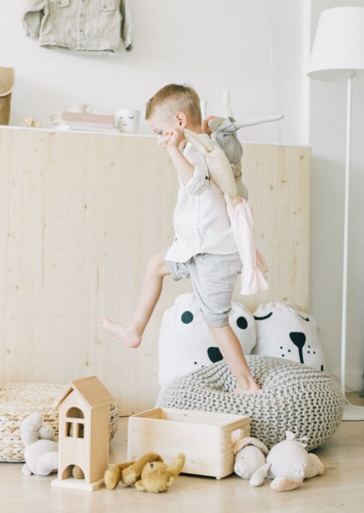 pexels-leeloothefirst-5029343 Ein kleiner Junge springt barfuß von einem grauen Strick-Sitzsack inmitten seines Spielzeugs und hält sich eine Stoffpuppe über die Schulter.