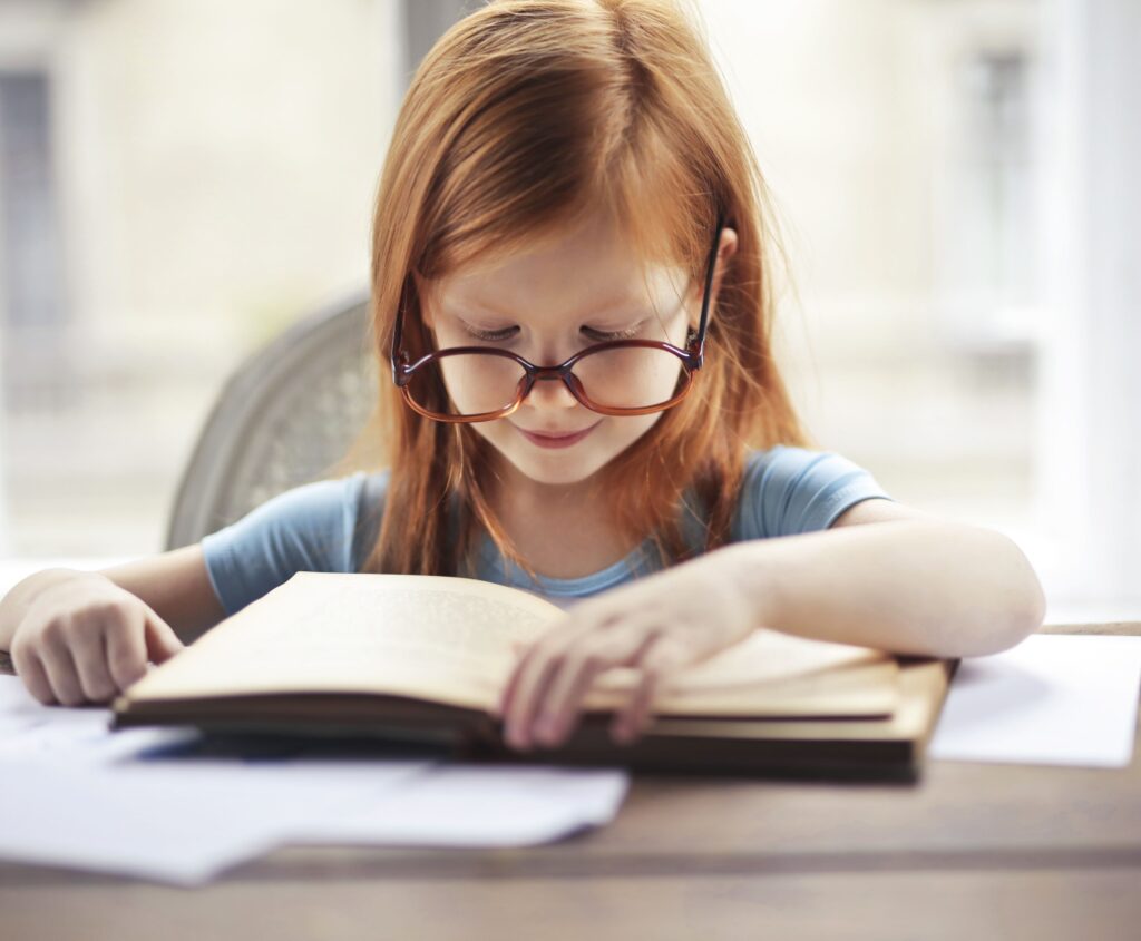 A young girl with glasses intently reads a book indoors, embodying the essence of learning and discovery.