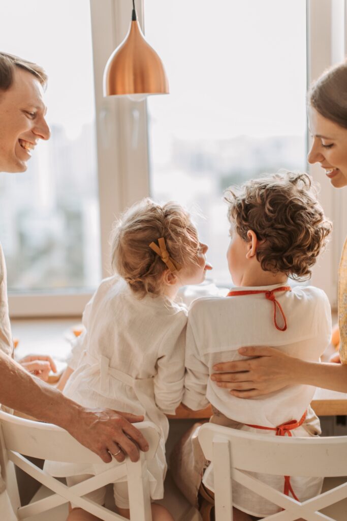 Eine Familie in der Küche, bei der ein kleines Mädchen mit einer Haarschleife scherzhaft die Zunge herausstreckt und ihren Bruder anschaut, während die Eltern lächelnd daneben stehen.